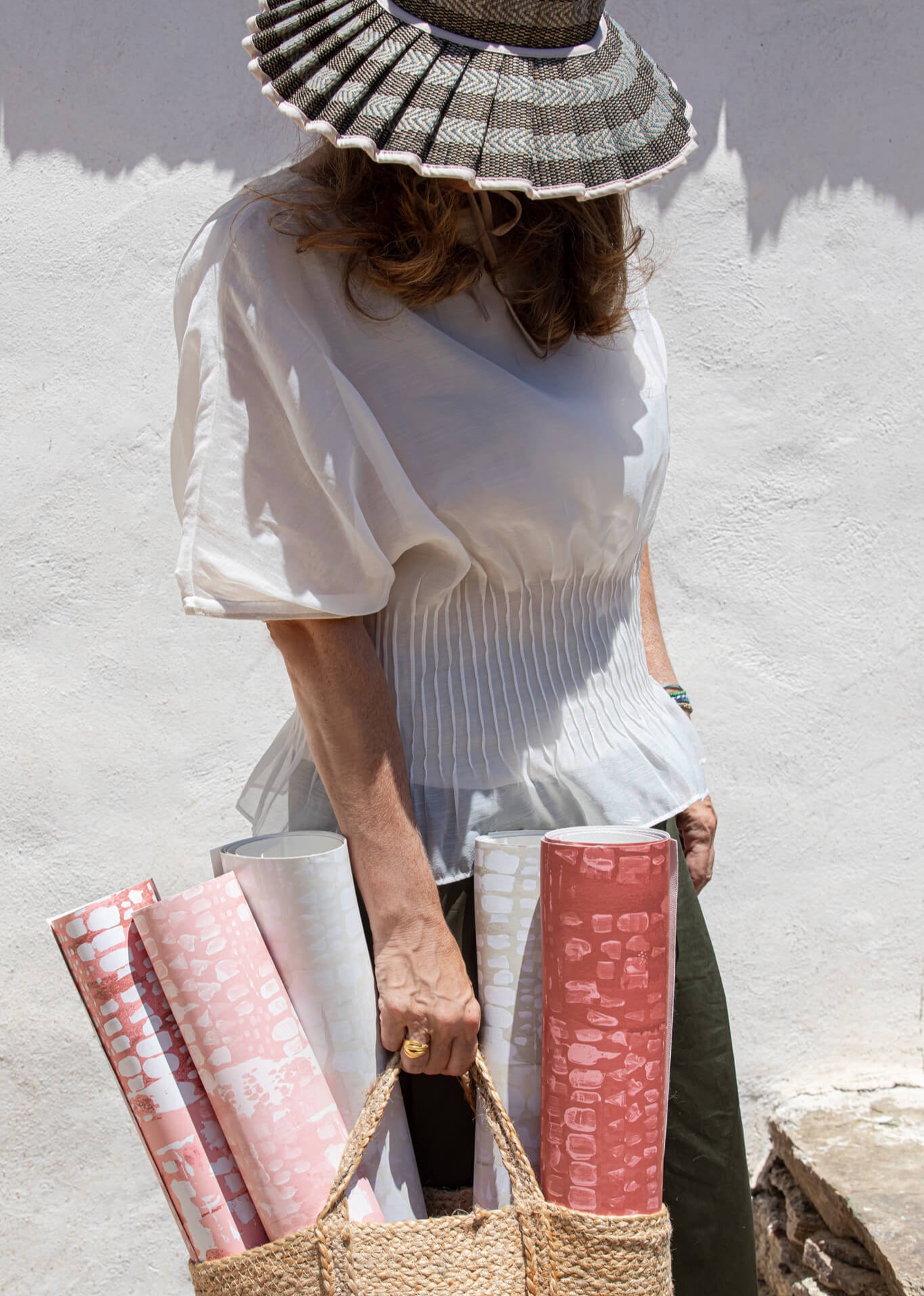 Woman holding rolled-up pink fabric and a woven bag against a white wall.