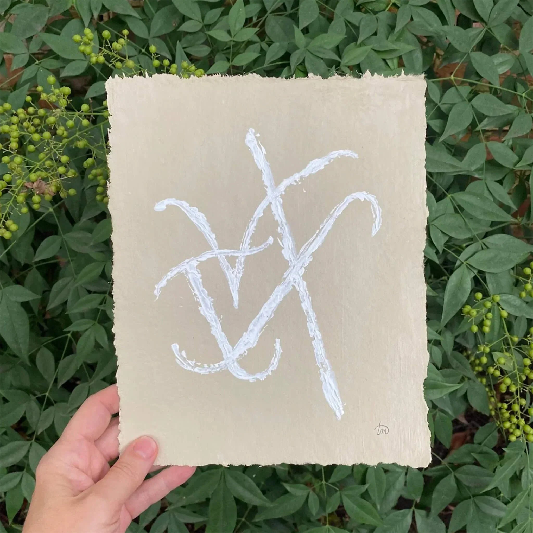 Custom Grammata painting by Richmond artist Theodora Miller on beige handmade cotton rag paper with deckled edges, featuring intertwined Greek initials in textured white paint, held against a backdrop of green foliage.